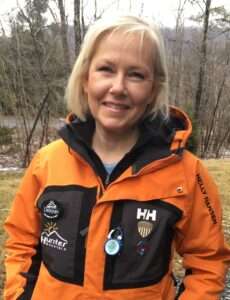A woman with blonde hair smiles outdoors, wearing an orange and black Helly Hansen jacket with various patches and pins. Trees and a river are visible in the blurred background.