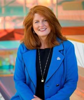 A woman with long red hair, wearing a bright blue blazer over a black top, smiles while sitting indoors. The colorful, blurred background suggests a modern building or office space.