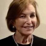 An older woman with light brown hair, wearing a black top, a pearl necklace, and pearl earrings, smiles while facing the camera against a plain, light-colored background.