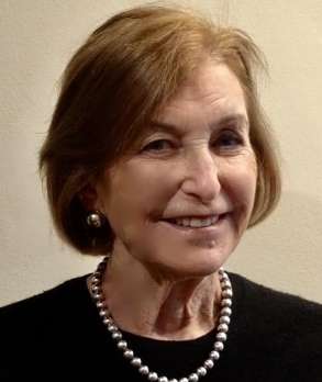An older woman with light brown hair, wearing a black top, a pearl necklace, and pearl earrings, smiles while facing the camera against a plain, light-colored background.