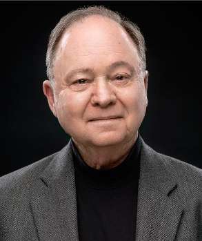 A man with short gray hair, wearing a gray blazer and a black turtleneck, smiles softly against a dark background.