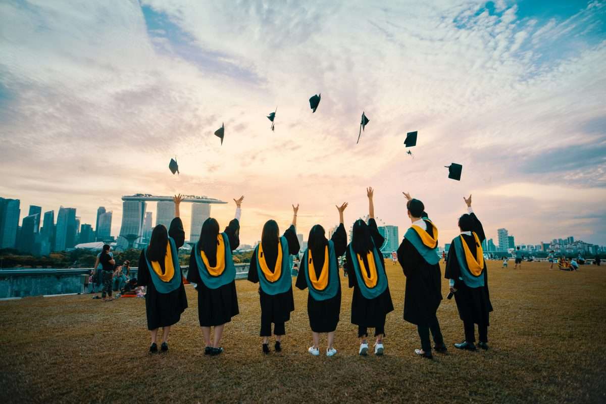 A group of graduates in caps and gowns stand outdoors, tossing their caps into the air, with a city skyline and dramatic cloudy sky in the background.