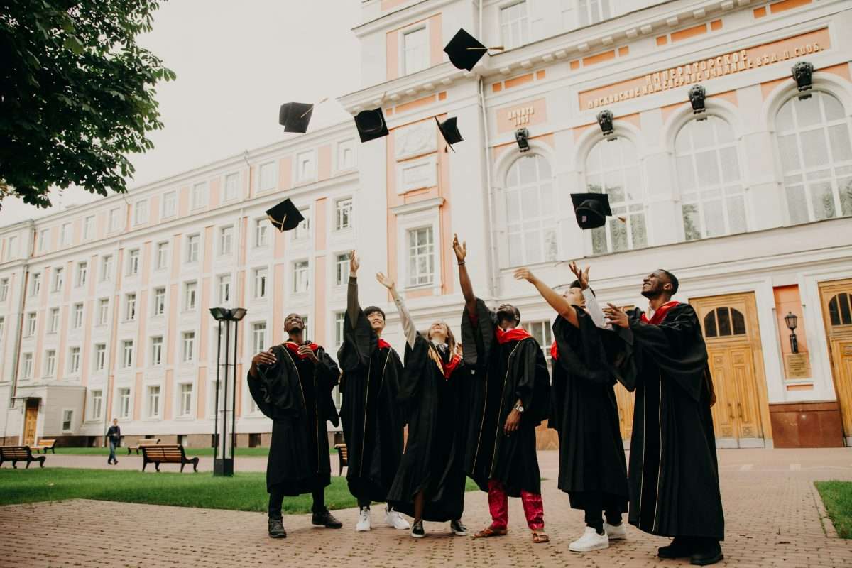 Six graduates in black gowns and red stoles stand outside a large building, joyfully tossing their caps into the air. The scene captures a celebratory moment in front of a university or academic institution.