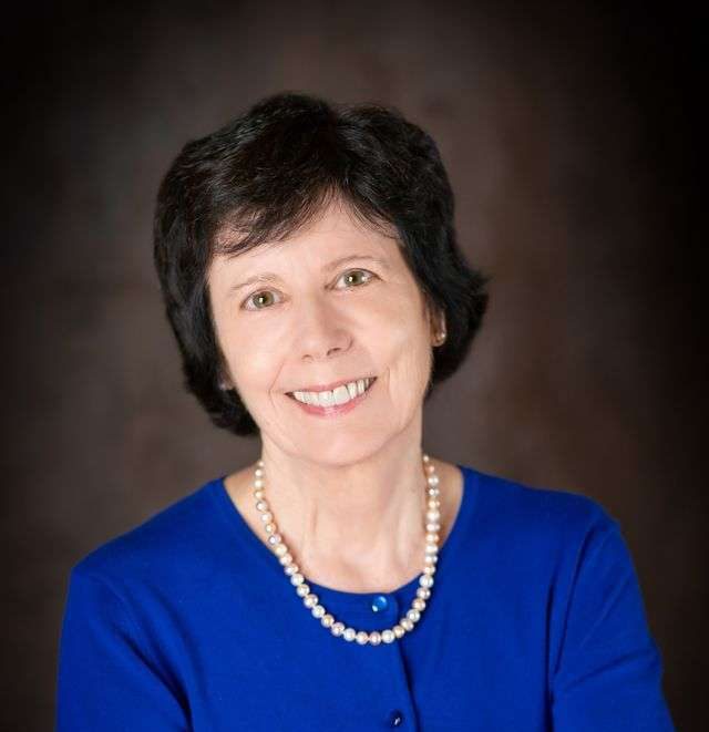 A woman with short dark hair, wearing a royal blue cardigan, a pearl necklace, and a gold ring, smiles with her arms crossed in front of a dark brown background.