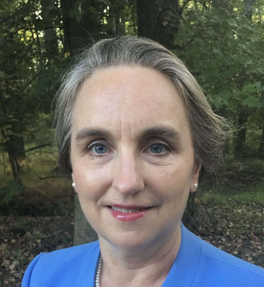 A woman with light skin, blue eyes, and grayish hair pulled back smiles gently. She wears a blue blazer and a pearl necklace, standing outdoors with green trees and foliage in the background.
