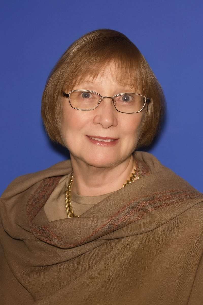 A woman with short light brown hair and glasses, wearing a beige shawl and a gold necklace, smiles in front of a solid blue background.