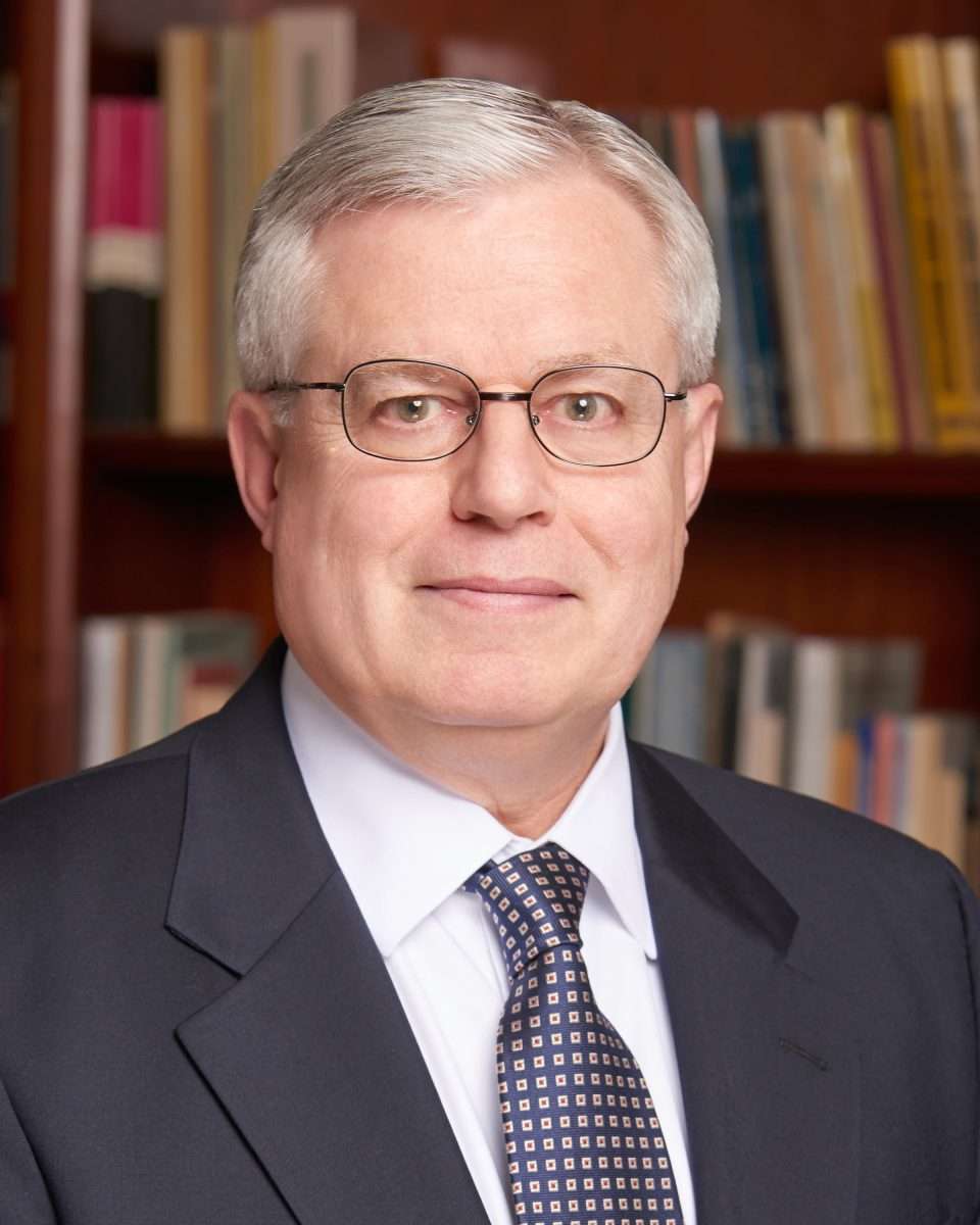 A man with short gray hair and glasses wearing a dark suit, white shirt, and patterned tie stands in front of a bookshelf filled with books.