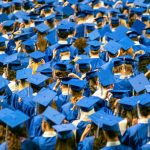 A large group of graduates wearing blue caps and gowns sit close together at a graduation ceremony, seen from behind. Many are adjusting their caps, and the crowd fills the frame.